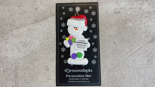 Ornament of white snowman in red hat holding various medications and a medical chart, shown on backing card and Christmas tree.