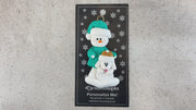 Ornament of snowman in green shirt and hat with white dog in Christmas hat, shown on backing card and Christmas tree.