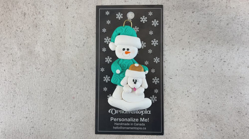 Ornament of snowman in green shirt and hat with white dog in Christmas hat, shown on backing card and Christmas tree.