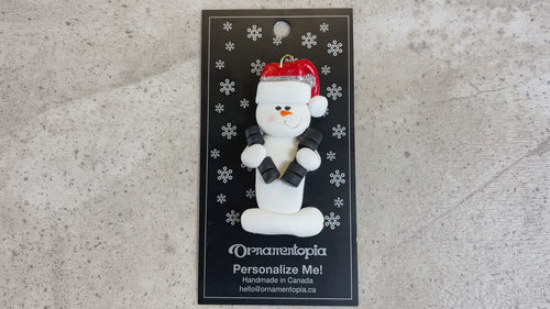 Ornament of white snowman in red hat carrying two dumbbells, shown on backing card and Christmas tree.