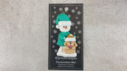 Ornament of snowman in green shirt and hat with tan dog in Christmas hat, shown on backing card and Christmas tree.
