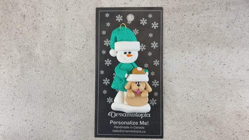 Ornament of snowman in green shirt and hat with tan dog in Christmas hat, shown on backing card and Christmas tree.