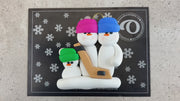 Family ornament with 3 white snowmen in bright hats, holding a hockey stick, shown on backing card and Christmas tree.