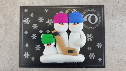 Family ornament with 3 white snowmen in bright hats, holding a hockey stick, shown on backing card and Christmas tree.