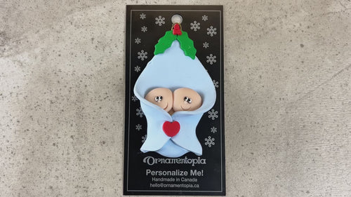 Ornament with twin infant heads poking out of a blue baby blanket, shown on backing card and Christmas tree.