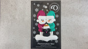 Ornament of snowman couple in red and green shirts and hats with black dog in Christmas hat, shown on backing card and Christmas tree.