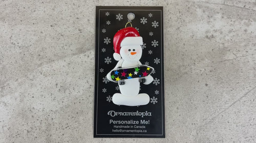 Ornament of white snowman in red hat holding a vibrant skateboard, shown on backing card and Christmas tree.