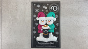 Ornament of snowman couple in red and green shirts and hats with a black cat in Christmas hat, shown on backing card and Christmas tree.
