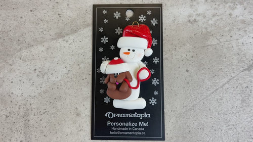 Ornament of white snowman in red hat holding a brown dog on a leash, shown on backing card and Christmas tree.