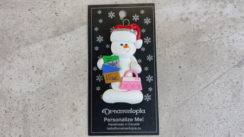 Ornament of white snowman in red hat holding a purse and various bank cards, shown on backing card and Christmas tree.