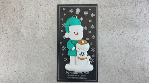 Ornament of snowman in a green shirt and hat with white cat in Christmas hat, shown on backing card and Christmas tree.