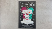 Ornament of snowman couple in red and green shirts and hats with a white cat in Christmas hat, shown on backing card and Christmas tree.