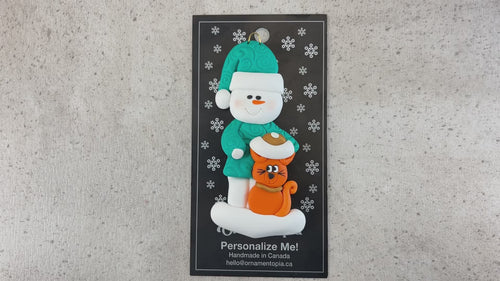 Ornament of snowman in a green shirt and hat with orange cat in Christmas hat, shown on backing card and Christmas tree.