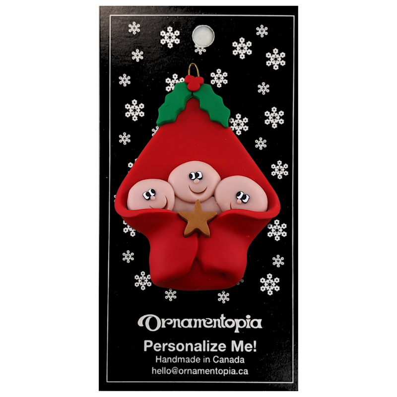 Ornament with triplet infant heads poking out of a red baby blanket, on display backing card.