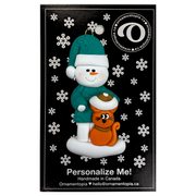 Ornament of snowman in a green shirt and hat with orange cat in Christmas hat, on display backing card.
