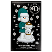 Ornament of snowman in a green shirt and hat with white cat in Christmas hat, on display backing card.