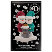 Ornament of snowman couple in red and green shirts and hats with grey cat in Christmas hat, on a display backing card.