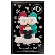 Ornament of snowman couple in red and green shirts and hats with black dog in Christmas hat, on display backing card.