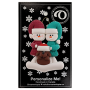 Ornament of snowman couple in red and green shirts and hats with brown dog in Christmas hat, on display backing card.