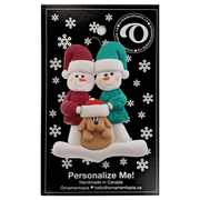Ornament of snowman couple in red and green shirts and hats with tan dog in Christmas hat, on display backing card.