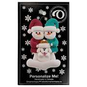 Ornament of snowman couple in red and green shirts and hats with white dog in Christmas hat, on display backing card.