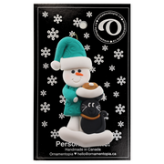 Ornament of snowman in a green shirt and hat with black cat in Christmas hat, on display backing card.