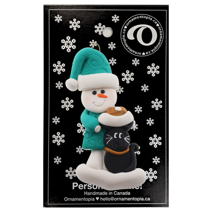 Ornament of snowman in a green shirt and hat with black cat in Christmas hat, on display backing card.