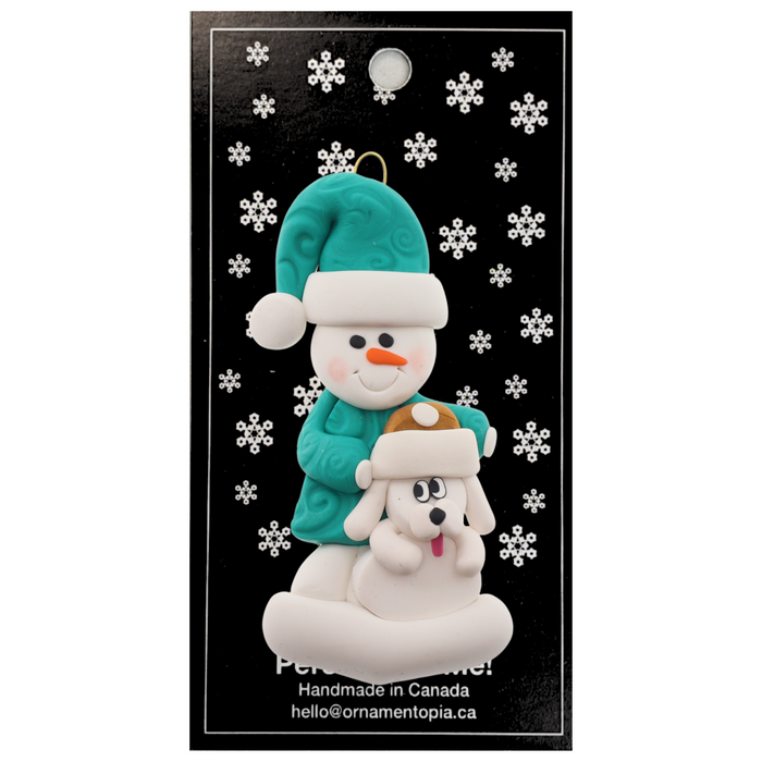 Ornament of snowman in green shirt and hat with white dog in Christmas hat, on display backing card.